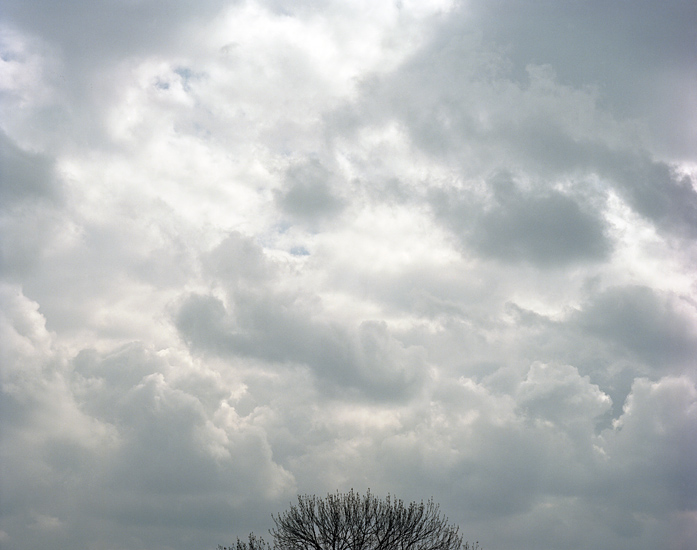 Foto einer dichten Wolkendecke mit den laublosen Spitzen eines Baumes am unteren Bildrand. Das Foto ist Teil des Projektes -Vollendete Gegenwart- des Kölner Fotografen Marcel Wurm.